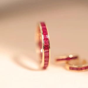Könnte beinhalten: Ein goldener Ring mit einem Band aus quadratisch geschliffenen, leuchtend roten Edelsteinen. Der Ring ist scharf abgebildet, mit zwei weiteren Ringen leicht unscharf im Hintergrund. Die Edelsteine sind gleichmäßig angeordnet.