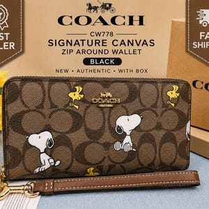 May include: A brown Coach signature canvas zip-around wallet featuring a repeating Snoopy and Woodstock print. The wallet includes a detachable brown leather wrist strap with gold-tone hardware, a gold Coach logo emblem, and a zipper closure.