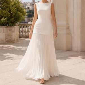 May include: A floor-length white dress featuring a sleeveless bodice with a boat neckline and a fitted waist. The lower portion of the dress consists of a long, flowing skirt with vertical pleats that extend from the waist to the hemline.