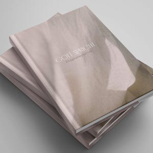 Könnte beinhalten: Ein Stapel Journale mit einem weichen, beigefarbenen Einband, der ein dezentes, abstraktes Licht- und Schattenmuster aufweist. Auf dem Vorderdeckel steht in weißer Serifenschrift "GOTT SPRICHT", gefolgt von dem kleineren Text "Prophetiejournal".