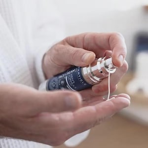 Op de afbeelding: Een blauwe en zilveren gezichtscr&egrave;mefles met de tekst "FACE CREAM" en "ARTICLE MEN'S FORMULA". De cr&egrave;me wordt op een hand aangebracht. Het product is voor huidverzorging.