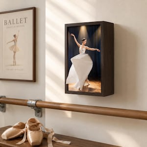 May include: A rectangular dark wood shadow box frame containing a backlit photograph of a ballerina in a white dress performing on a stage. To the left, a framed print titled BALLET GRACE IN MOTION hangs above a wooden barre and ballet slippers.