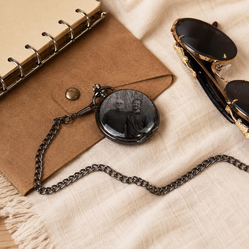 May include: A round, black metal pocket watch featuring a circular black and white photograph of two people on its cover. The watch is attached to a matching black metal chain that extends across the frame, resting near a pair of dark sunglasses.