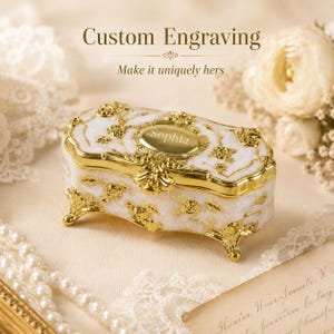 May include: A rectangular jewelry box with a white marbled finish and gold-toned metal accents featuring floral rose motifs. The lid displays an oval gold plaque engraved with the name Sophia. The box stands on four ornate gold-toned feet.