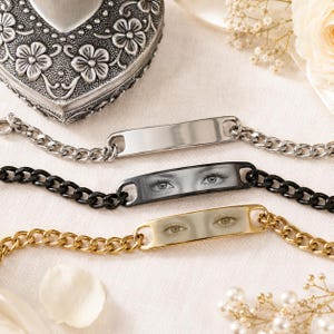 Puede incluir: Tres pulseras de cadena con placas metálicas rectangulares. La placa superior es de tono plateado pulido, la central es negra con un estampado de ojos en escala de grises y la inferior es de tono dorado con un estampado de ojos en sepia.