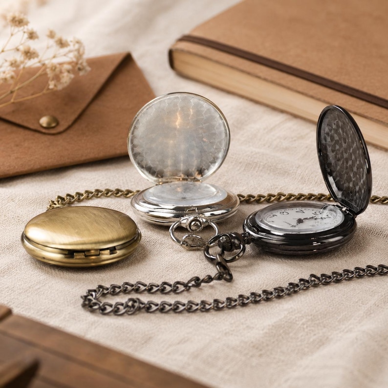May include: Three pocket watches with metal chains. One is a closed antique brass-toned watch. Another is an open silver-toned watch with a reflective interior. The third is an open black watch with a white clock face featuring black numerals.