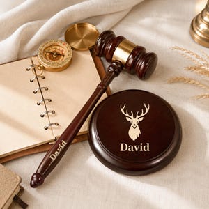 Può includere: Un martelletto in legno marrone scuro e una base circolare abbinata con la sagoma di una testa di cervo incisa e il nome David. Anche il manico del martelletto riporta inciso il nome David. Una bussola in ottone poggia su un taccuino a spirale.