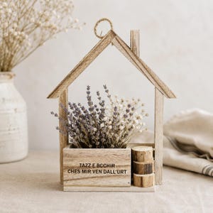 May include: A house-shaped wooden frame containing a rectangular planter box filled with dried lavender and white flowers. The front of the box features the text TAZZ E BCCHIR CHES MIR VEN DALL'URT and is decorated with three small stacked wooden logs.