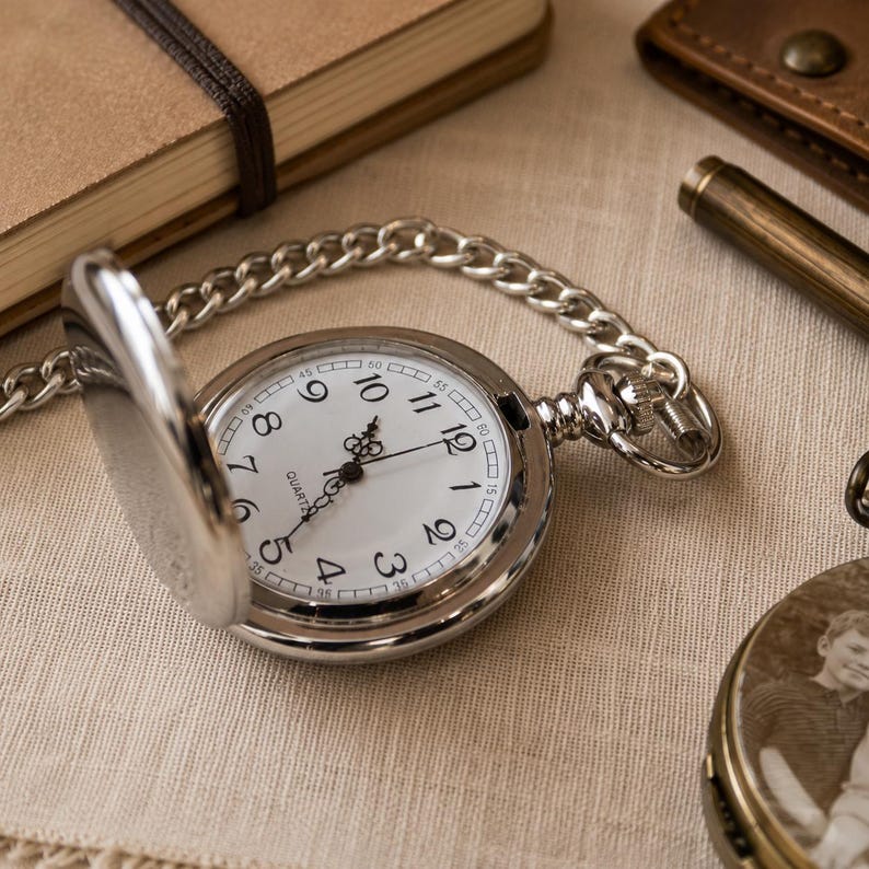 May include: A silver-toned pocket watch with an open cover, featuring a white dial with black Arabic numerals, ornate black hands, and the word QUARTZ printed on the face. The watch is attached to a matching silver-toned metal link chain.