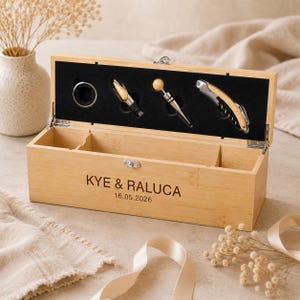 May include: A rectangular bamboo wine box with a hinged lid, featuring the engraved text KYE & RALUCA 16.05.2026. The interior lid contains a black foam insert holding a metal drip ring, a wine pourer, a bottle stopper, and a corkscrew tool.