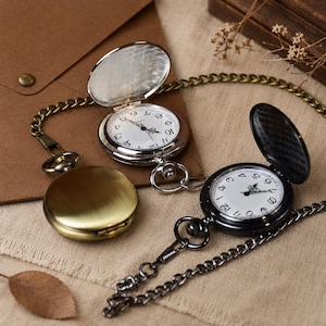 May include: Three pocket watches with metal cases in silver, brass, and black finishes. Two watches are open, revealing white faces with black Arabic numerals and hands. Each watch features a matching metal chain attached to the top crown.