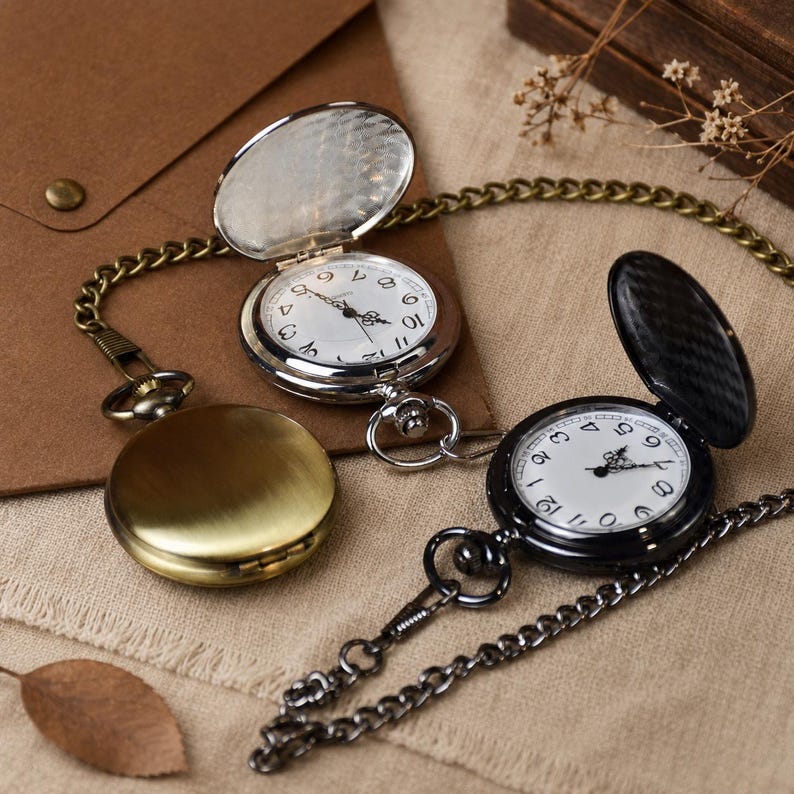 May include: Three pocket watches with metal cases in silver, brass, and black finishes. Two watches are open, revealing white faces with black Arabic numerals and hands. Each watch features a matching metal chain attached to the top crown.