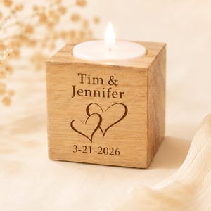 May include: A square wooden tea light candle holder featuring the engraved names Tim & Jennifer, two heart outlines, and the date 3-21-2026. A white tea light candle with a lit flame sits in the circular cutout on the top surface of the block.