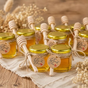 Könnte beinhalten: Eine Sammlung kleiner sechseckiger Gläser gefüllt mit goldenem Honig, jeweils mit einem goldenen Metalldeckel. Ein hölzerner Honiglöffel ist mit einer Schnur an jedem Glas befestigt, zusammen mit einem runden Anhänger mit Thank You und einem roten Herz.