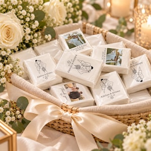 May include: A woven basket filled with square white wedding favor packets. Some packets feature a black line drawing of a couple with the text Adeline & Jérémy 27.06.26, while others display a color photograph of a couple and the text Patrick & Katie 26.06.26.