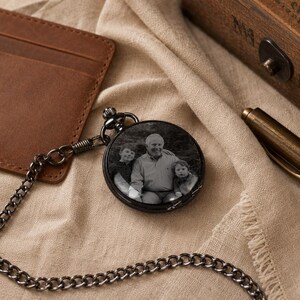 May include: A round black pocket watch featuring a custom black and white photograph on its cover. The image displays an adult and two children posing together. The watch is attached to a matching black metal chain, creating a classic timepiece.