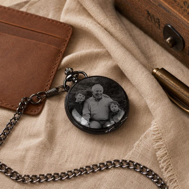 May include: A round black pocket watch featuring a custom black and white photograph on its cover. The image displays an adult and two children posing together. The watch is attached to a matching black metal chain, creating a classic timepiece.