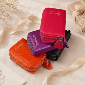 Op de afbeelding: Drie rechthoekige, getextureerde lederen etuis met ritssluiting in fel oranje, paars en roze, met een zwarte eronder. Elk etui is voorzien van een zilverkleurige dubbele ritssluiting en is gepersonaliseerd met de namen Gerard, Sandra en Lavande.