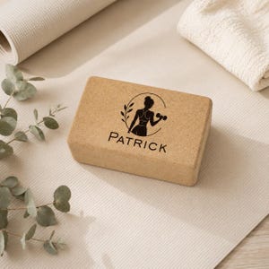 May include: A rectangular cork yoga block featuring a black graphic print of a person holding a dumbbell inside a circular frame with a leaf branch. The name PATRICK is printed in black capital letters directly below the graphic illustration.