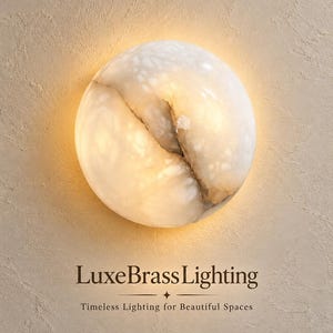 May include: A circular wall sconce made of translucent white stone with natural veining, emitting a warm glow. Below the light, the text reads: LuxeBrassLighting, Timeless Lighting for Beautiful Spaces.