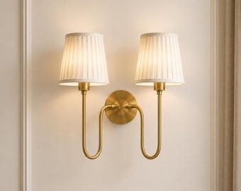 May include: A double wall sconce featuring two off-white pleated fabric lampshades mounted on a central circular brass base. The fixture has two curved brass arms extending outward, each supporting a single light source directed downward.