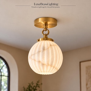May include: A flush mount ceiling light featuring a round, ribbed white stone globe shade with natural veining, suspended from a circular brushed brass canopy. The text LuxeBrassLighting Timeless lighting for beautiful spaces is visible.