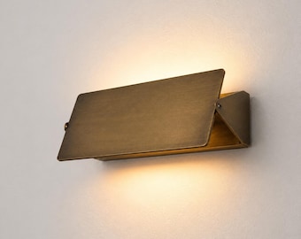May include: A rectangular wall sconce with a brushed brass finish. The fixture features a flat, angled front panel that directs light both upward and downward, creating a warm, soft glow against the wall surface behind the metal structure.