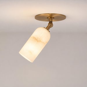 May include: A ceiling-mounted spotlight featuring a cylindrical, translucent white stone shade with natural veining. The fixture includes a circular brass base and an adjustable brass swivel joint that allows the light to be angled as needed.