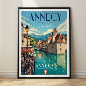 Puede incluir: Un póster de viaje enmarcado de Annecy, Francia, que muestra una escena de canal con edificios de piedra, una bicicleta y montañas bajo un cielo azul. El texto dice ANNECY, HAUTE-SAVOIE, FRANCE, La Venise des Alpes, e incluye un escudo rojo y blanco.