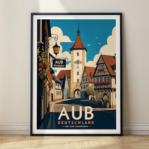 Op de afbeelding: Een ingelijste reisposter met een gestileerde illustratie van de stad Aub in Duitsland. De afbeelding toont een geplaveide straat die naar een hoge klokkentoren leidt, geflankeerd door vakwerkhuizen met rode daken, een hangende lantaarn en de tekst AUB DEUTSCHLAND - TOR ZUM GAUBÜNDEN -.