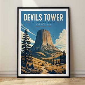 Op de afbeelding: Een ingelijste reisposter met het Devils Tower National Monument in Wyoming, VS. De illustratie toont de rotsformatie onder een blauwe lucht met wolken, een kronkelend pad en dennenbomen in een landschap met bruine en donkerblauwe tinten.