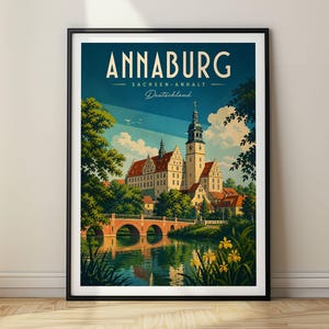 May include: A framed travel poster featuring the text ANNABURG SACHSEN-ANHALT Deutschland. The illustration depicts a historic castle with a tall tower, a red-roofed building, a stone bridge over a river, green trees, and yellow flowers.