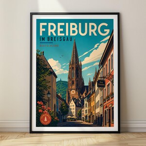 May include: A framed travel poster featuring a street view of Freiburg im Breisgau, Germany. The illustration shows a tall cathedral spire, colorful buildings, and a blue sky with clouds. The text reads FREIBURG IM BREISGAU DEUTSCHLAND.