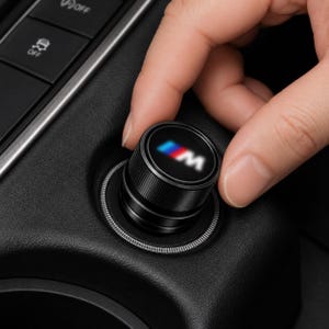 May include: A black cylindrical car interior control knob featuring a textured grip and a circular top surface displaying the M logo in blue, purple, and red colours. The knob is mounted on a black textured console panel with a silver trim ring.