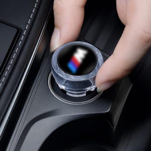 May include: A circular, clear plastic automotive accessory cap featuring a black center insert with a blurred M logo in blue, red, and white. The cap is designed to fit into a standard vehicle cigarette lighter or power outlet socket.