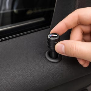 Puede incluir: Un pasador de bloqueo de puerta de coche cilíndrico negro con una parte superior circular que muestra el logotipo M en colores azul, morado y rojo. El pasador de metal tiene un acabado mate liso y está instalado en el panel interior negro.