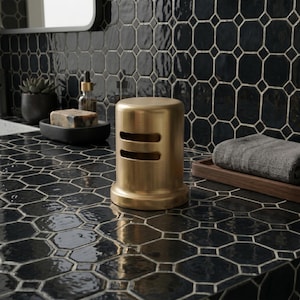 May include: A gold-colored, cylindrical soap dispenser with two horizontal slots. The dispenser sits on a black, geometric-patterned surface, with a matching tiled wall in the background. A rolled gray towel rests on a wooden tray.