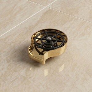 Pode incluir: Um tampão de garrafa decorativo dourado com um design de leme preto. O tampão tem uma base curva e retangular e um design de roda detalhado na parte superior. O tampão é feito de metal e assenta numa superfície clara.