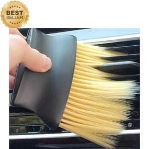 May include: A black car vent brush with light yellow bristles is being used to clean a car's air vents. The brush has a curved handle and is labeled "Best Seller" in a gold seal.