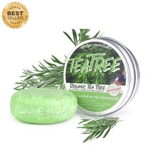May include: A green, round shampoo bar next to a metal tin with the words "Tea Tree" and "Organic Tea Tree" printed on it. The bar is labeled "Cruelty Free No Silicone Oil". The image also includes sprigs of green leaves.