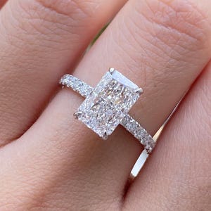 May include: A radiant-cut diamond engagement ring featuring a large rectangular center stone held by four prongs, set on a thin, polished metal band adorned with a row of small, round-cut pavé diamonds extending along the sides of the ring shank.