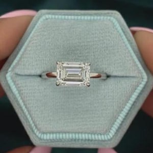 Può includere: Un anello d'argento con un diamante taglio smeraldo è presentato in un astuccio esagonale azzurro. L'anello è tenuto da una persona con lo smalto rosa, su uno sfondo verde acqua.