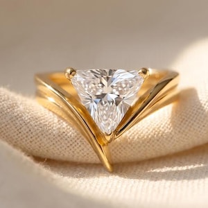 May include: A gold ring featuring a large, clear, trillion-cut gemstone set in a V-shaped band. The metal has a polished, reflective finish, and the triangular stone is faceted to catch the light, creating a bright, sparkling appearance.