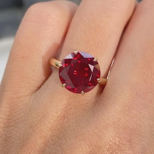 Pode incluir: Um anel de ouro com uma grande pedra preciosa vermelha escura, redonda e facetada. A pedra preciosa é o ponto focal, definida em uma banda de ouro simples e clássica. O anel é mostrado em uma mão.