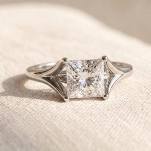 May include: A silver-toned ring featuring a central princess-cut clear gemstone held by four prongs. The band has a split-shank design that flares outward to meet the corners of the square-shaped stone, creating a geometric, open-work metal frame.