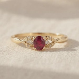 Peut inclure: Une bague en or ornée d'un rubis rouge central de taille ovale. L'anneau présente un motif délicat en forme de feuilles de chaque côté de la pierre, chacune agrémentée de petits diamants ronds et transparents incrustés dans les feuilles.