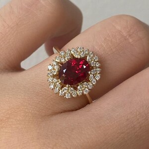 May include: A gold ring featuring a large, oval-cut, deep red gemstone surrounded by a halo of smaller, clear gemstones. The ring's band is thin and gold. The setting and band appear to be made of gold.