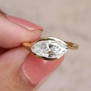 May include: A gold ring featuring a marquise-cut diamond. The diamond is set in a bezel setting, with the gold band curving around the stone. The ring is held up by a person's hand.