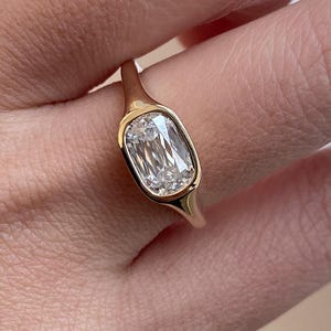 Op de afbeelding: Een gouden ring met een rechthoekige, heldere edelsteen. De ring heeft een eenvoudig, elegant ontwerp met een gladde, gepolijste band. De edelsteen is gezet in een bezel-zetting.