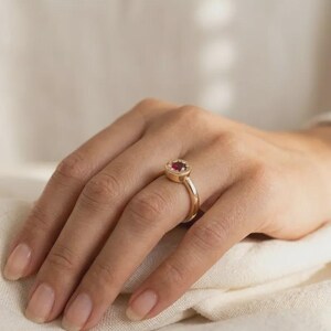May include: A gold ring featuring a round, deep red gemstone set in a circular bezel. The band is smooth and polished, showcasing a simple, clean design that highlights the vibrant color of the central stone and the warm tone of the metal.